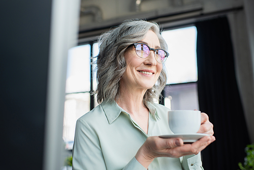 Happy grey haired businesswoman in eyeglasses holding cup and saucer in office
