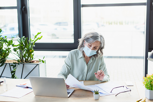 Mature businesswoman in medical mask working with documents near laptop in office