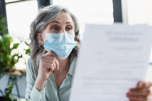 Grey haired businesswoman in medical mask looking at blurred document in office