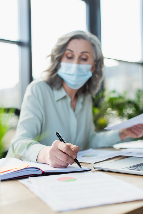 Blurred businesswoman in medical mask writing on notebook near laptop in office