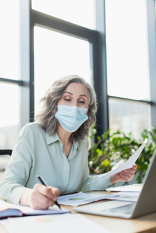 Grey haired businesswoman in medical mask holding paper and writing on notebook near laptop in office