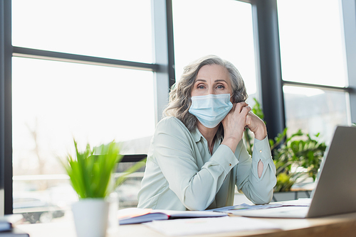 Mature businesswoman in medical mask  near laptop in office