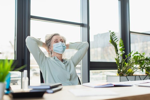 Mature businesswoman in medical mask sitting with closed eyes near notebooks in office