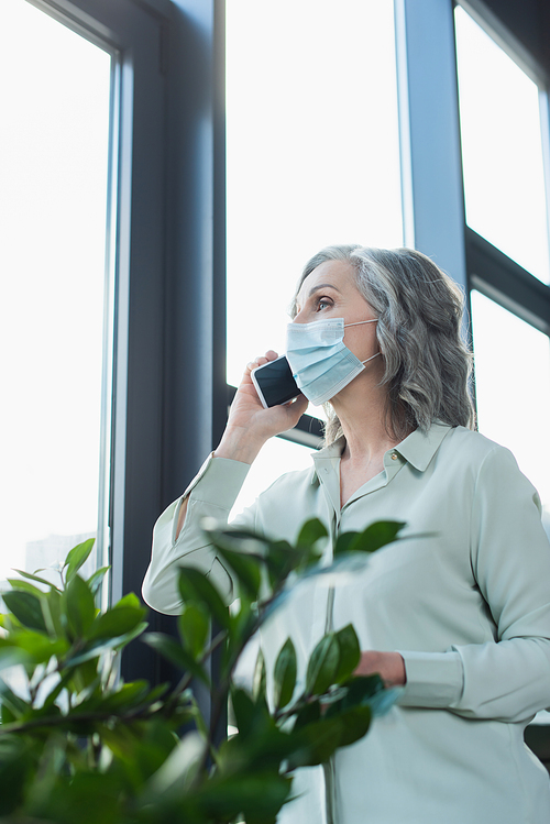 Mature businesswoman in medical mask talking on smartphone near plant and window in office