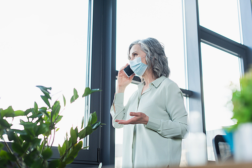 Mature businesswoman in protective mask talking on cellphone near window and plant in office