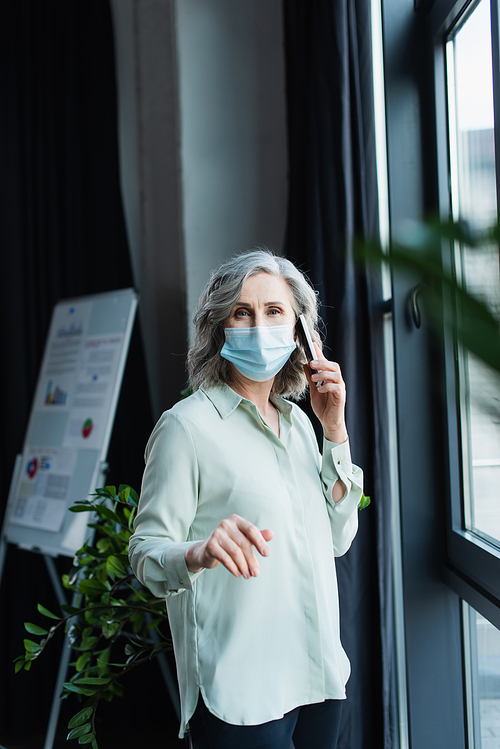 Mature businesswoman in medical mask talking on cellphone and  in office