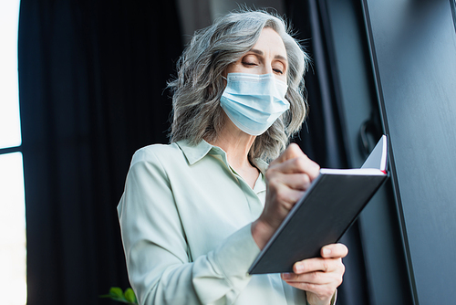 Low angle view of grey haired businesswoman in medical mask holding notebook in office