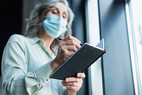 Low angle view of blurred businesswoman in medical mask writing on notebook in office