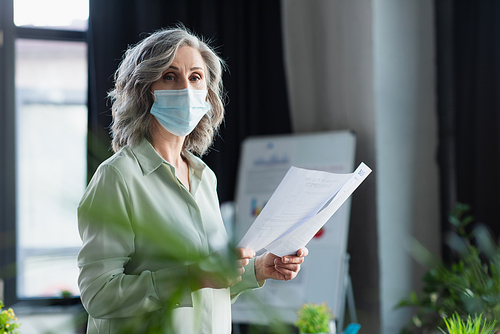 Grey haired businesswoman in medical mask holding documents and  in office