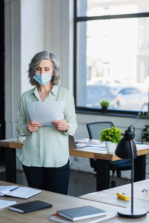Mature businesswoman in medical mask holding papers near notebooks on table