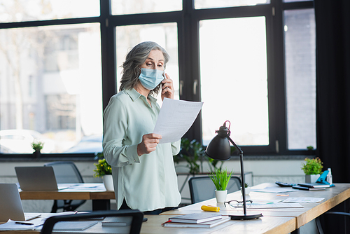 Mature businesswoman in medical mask talking on smartphone and holding document in office