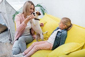 Cheerful woman playing with jack russell terrier near daughter on couch