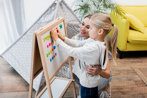 Smiling mother hugging kid near magnetic easel and blurred tent at home