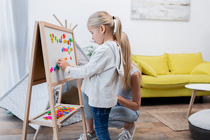 Girl playing with colorful magnets on easel near mom at home