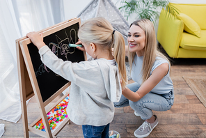 Positive woman looking at daughter drawing on chalkboard at home