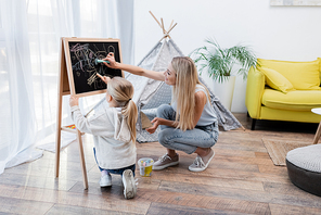 Mom and girl drawing on chalkboard near teepee at home