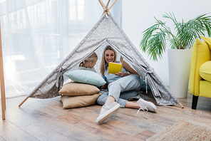 Mother reading book near child in teepee at home