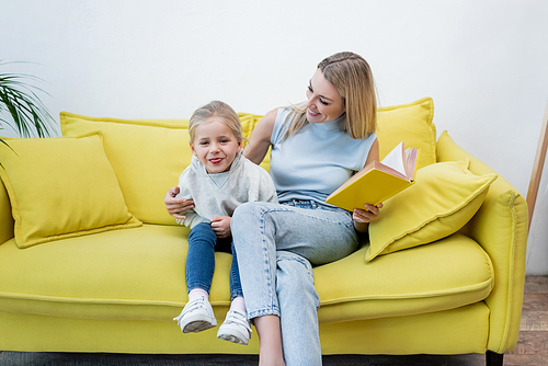 Kid sticking out tongue near mom with book on couch at home