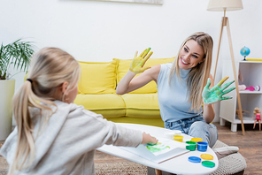 Smiling woman showing hands in paint near blurred kid at home