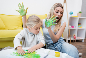 Shocked mother holding hands in paint near kid at home