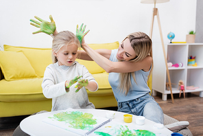 Positive woman holding hands in paint near daughter and sketchbook on coffee table at home