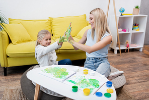 Smiling kid and mother showing hands in paint near sketchbook on coffee table in living room