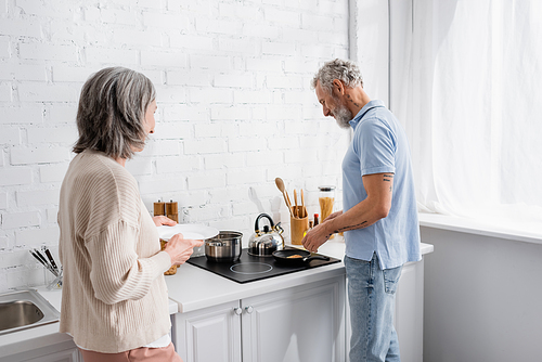 Mature man cooking pancake near wife with plate in kitchen