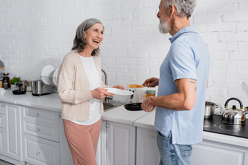 Positive woman holding plate near husband with frying pan and pancake in kitchen