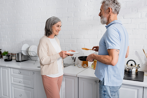 Woman holding plate near husband with pancake and frying pan in kitchen