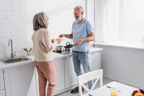 Happy mature man holding frying pan and pancake near wife with plate in kitchen