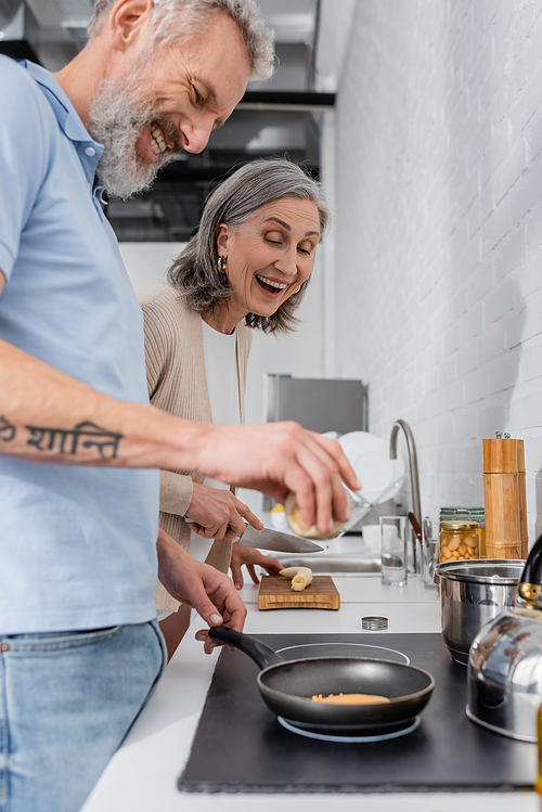 Cheerful woman holding knife near husband cooking pancake in kitchen
