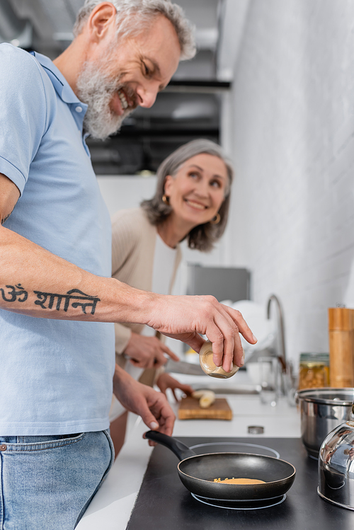 Smiling man holding spice while cooking pancake near blurred wife in kitchen