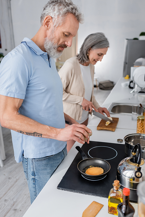 Side view of man seasoning pancake near blurred wife cutting banana in kitchen