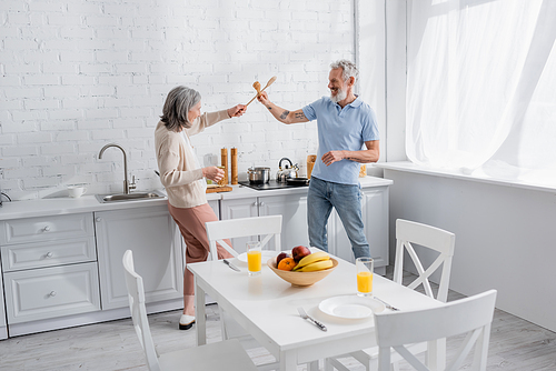 Side view of smiling mature couple fighting with spatulas in kitchen