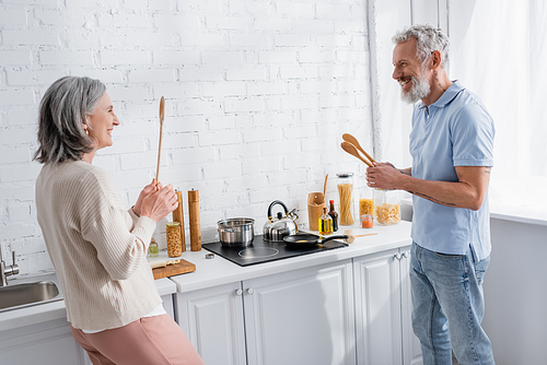 Happy mature couple holding spoons near stove in kitchen
