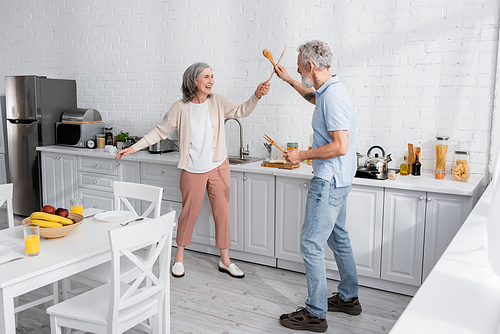 Happy mature woman fighting with wooden spatulas in kitchen