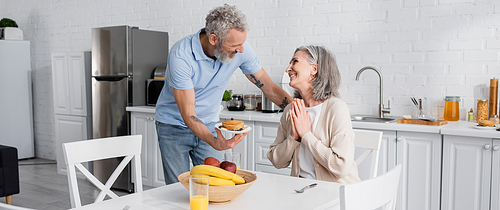 Smiling woman showing please gesture near mature husband with pancakes in kitchen, banner