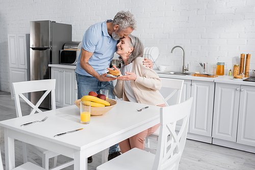Mature man kissing wife with pancakes near orange juice and fruits in kitchen