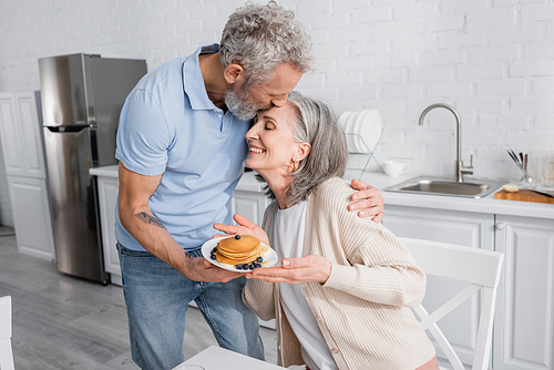 Mature man kissing smiling wife with tasty pancakes in kitchen