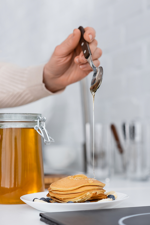 Cropped view of woman pouring honey on pancakes with berries in kitchen