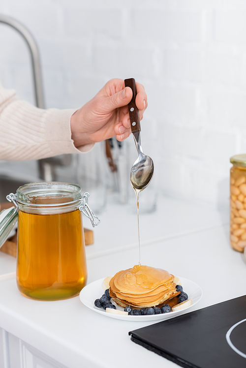 Cropped view of woman pouring honey on pancakes with fruits in kitchen
