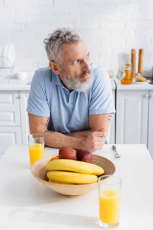 Mature tattooed man looking away near fruits and orange juice in kitchen