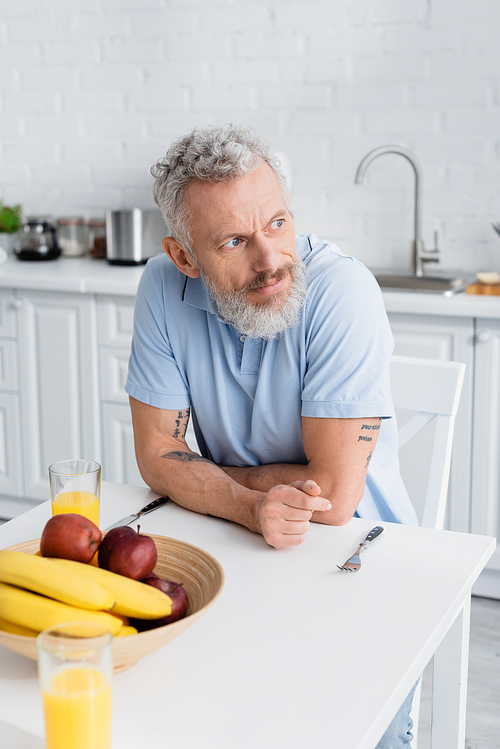 Middle aged man sitting near fruits and orange juice in kitchen