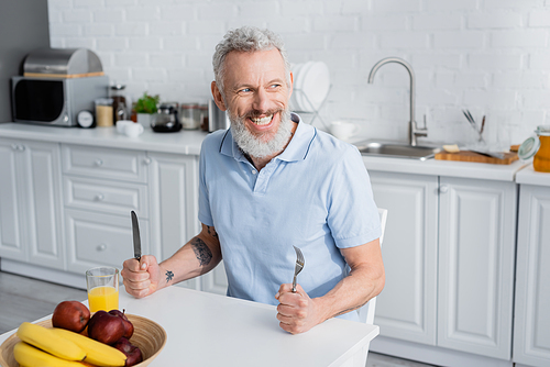 Smiling mature man holding cutlery near orange juice and fruits in kitchen