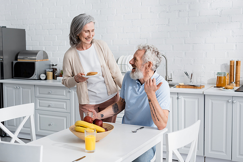 Smiling woman holding pancakes near cheerful husband and fruits in kitchen