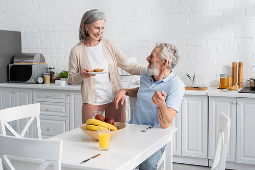 Smiling woman holding pancakes near husband and fruits in kitchen