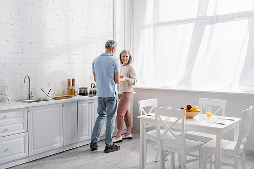 Mature man holding hand of smiling wife near worktop in kitchen
