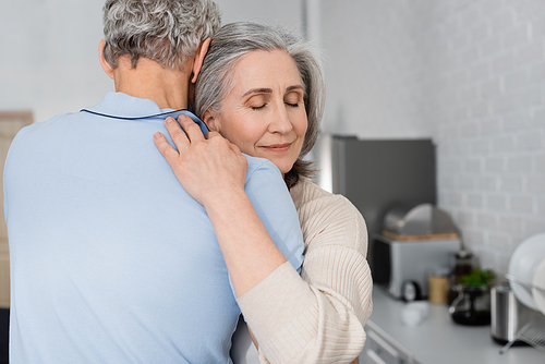 Mature woman with closed eyes embracing husband in kitchen