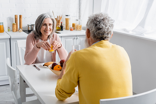 Positive woman holding glass of orange juice near fruits and blurred husband at home