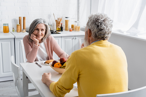 Mature woman looking at blurred husband near breakfast in kitchen
